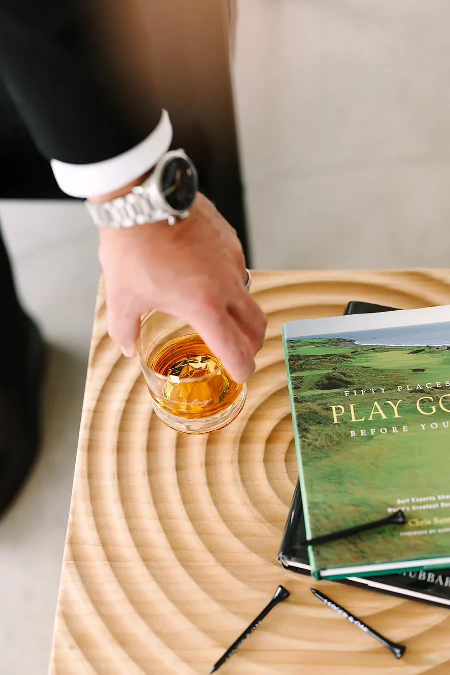 A hand with a silver watch holding a rocks glass over a carved wooden table beside a golf book